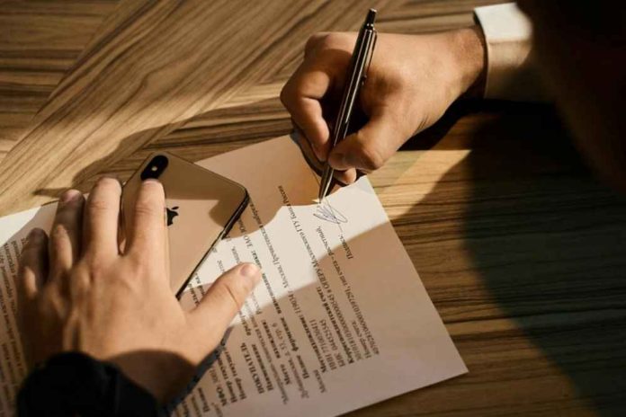 A person signing a document on the wooden table while holding a phone